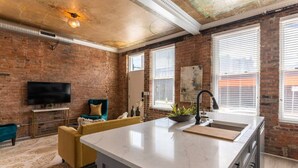 A modern kitchen with a marble countertop, a sink, and a built-in oven. There is a brick wall and a large window with blinds.