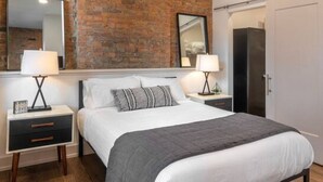 Une chambre moderne avec un mur en briques, un lit recouvert de linge de lit blanc et d’un plaid gris, deux tables de chevet avec des lampes et un ventilateur de plafond.