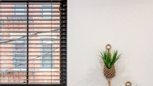 Une fenêtre avec des stores horizontaux et trois pots à plantes en macramé, fixés au mur et contenant des plantes vertes.