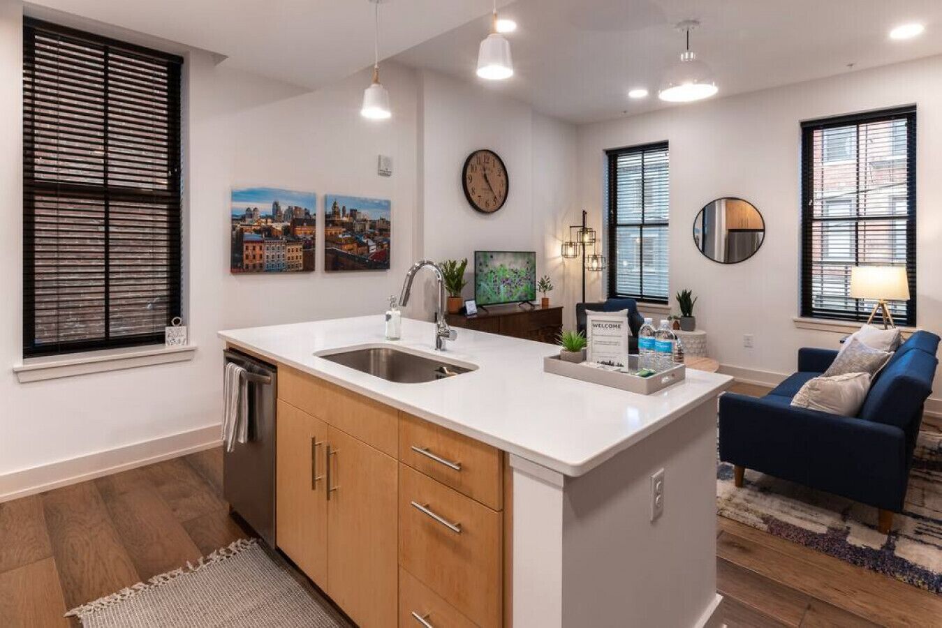 A modern kitchen with a central island, double sinks, and wooden cabinets. There are two windows with blinds, a clock on the wall, and a cityscape painting.