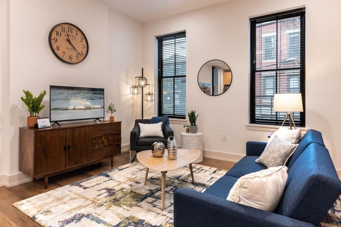 Una sala de estar con sofá, mesa de centro, televisor, reloj y dos ventanas con persianas.