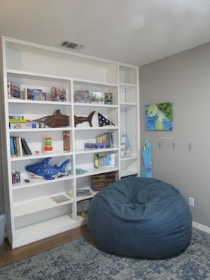 Ein Zimmer mit einem weißen Bücherregal, gefüllt mit Büchern und Spielzeug, einem blauen Sitzsack und einem Bild an der Wand.