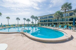 Una piscina con sillones de descanso y palmeras, rodeada por un edificio con varios pisos y balcones.
