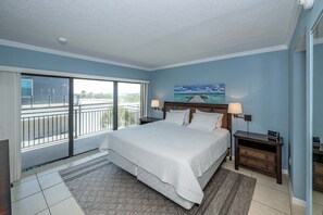 A bedroom with a large bed, two bedside tables, a view of the outdoors through a sliding door, and a painting on the wall.