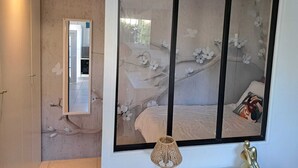 Un dormitorio con una ventana grande, una cama, una lámpara de noche y un espejo.