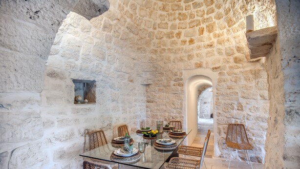 A stone-walled dining area with a glass table, wicker chairs, and a view of a courtyard. - Trullo La Succulenta - Le Case di Valentina (Ceglie Messapica)