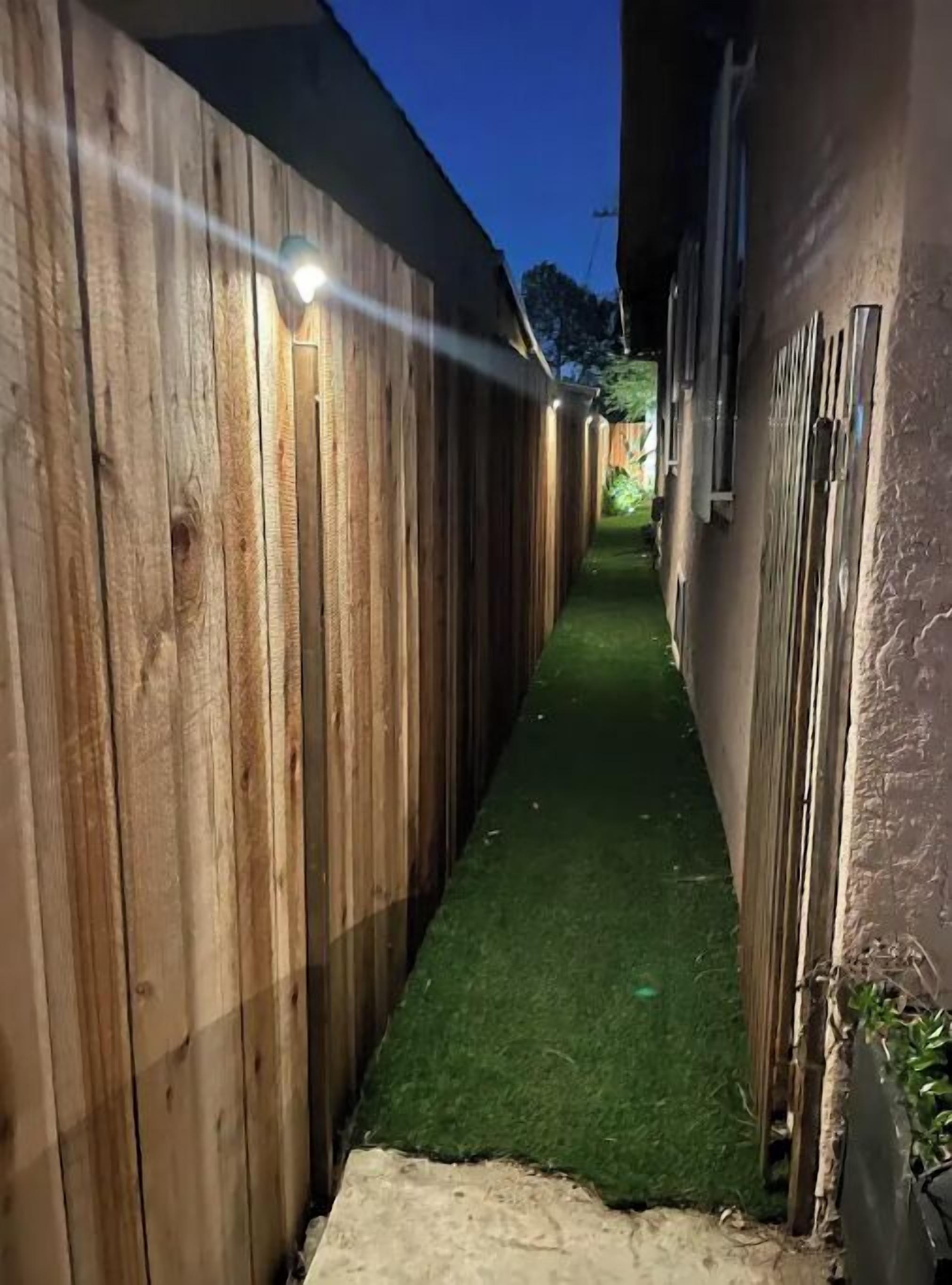 A narrow pathway flanked by wooden fences with artificial grass, leading to a building with a dark exterior.