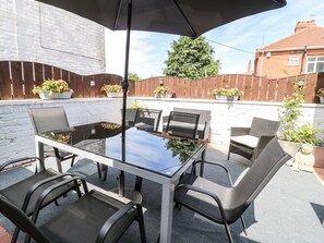 A patio with a glass-top table and six chairs, surrounded by potted plants and a wooden fence.