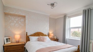 A bedroom with a wooden bed, two bedside tables with lamps, a window with curtains, and a wall with floral wallpaper. - Westfield (Bridlington)