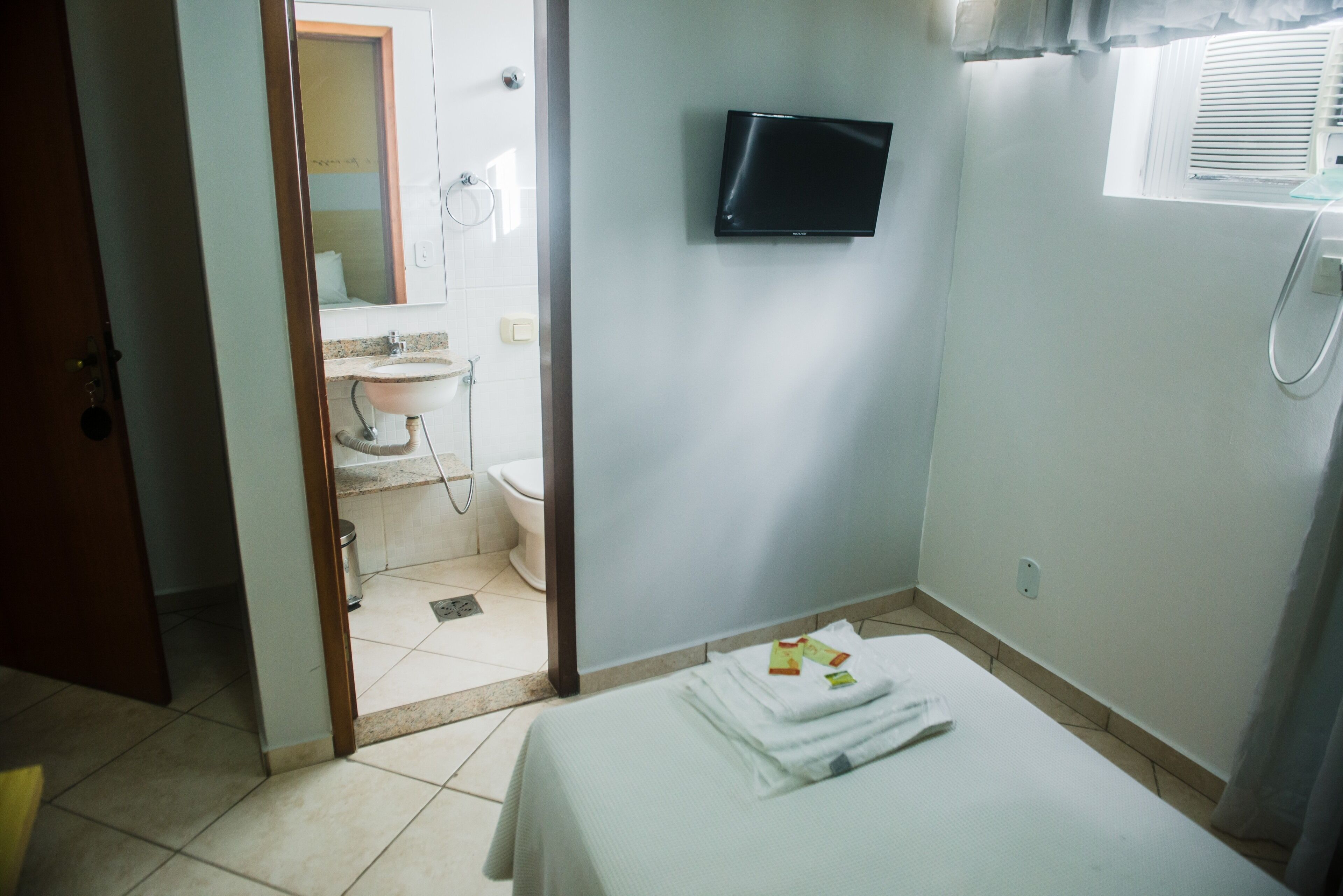 Ein Hotelzimmer mit Bett, Wandfernseher, Bad mit Waschbecken und Toilette sowie einem Fenster mit Jalousien.