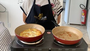 Una persona con delantal y gorro negro está de pie en una cocina con dos ollas sobre una estufa portátil. La cocina tiene una pared blanca con un cartel de ‘No Smoking’ y un cartel del ‘Minasdor Hotel’.