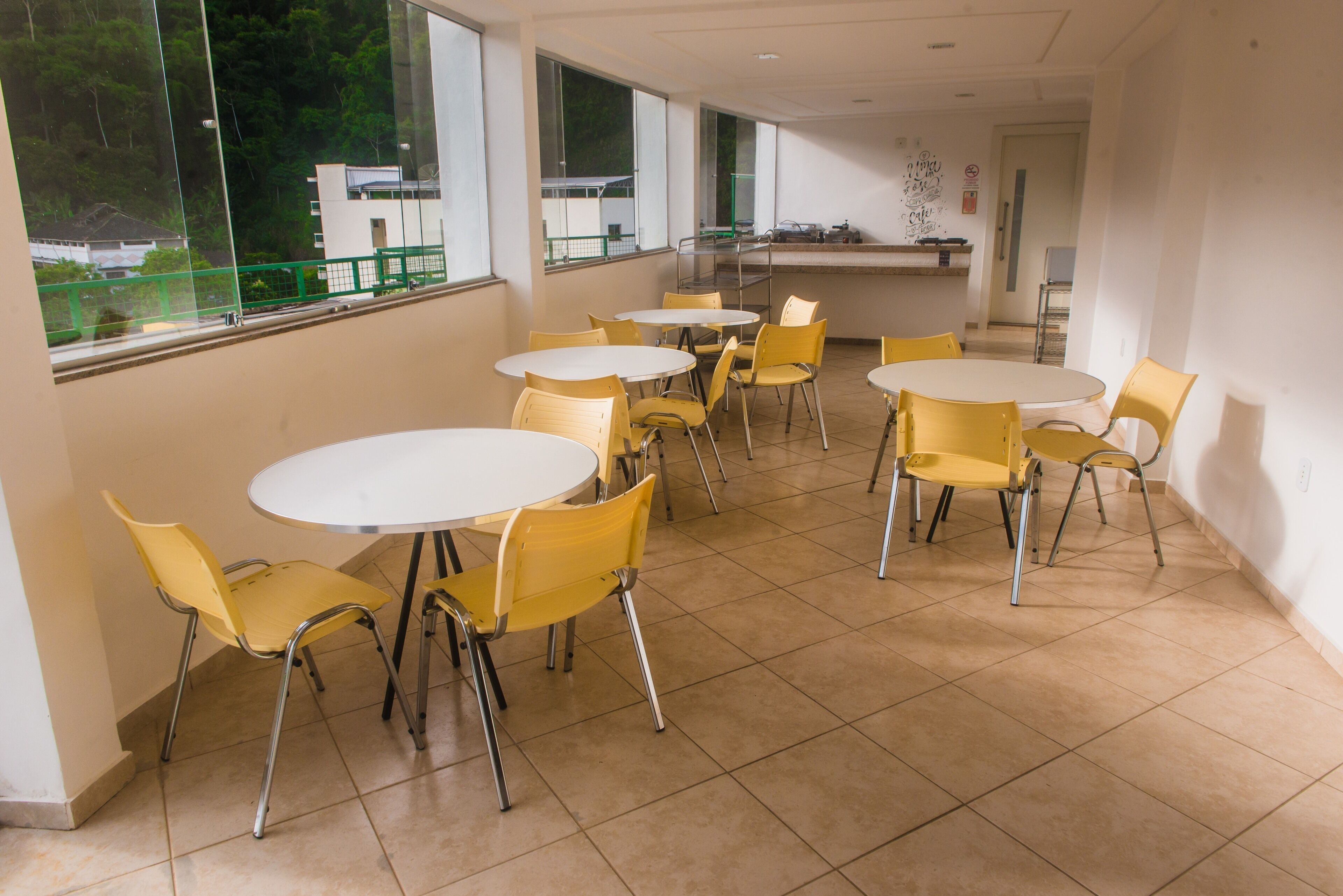 A dining area with round tables and yellow chairs, large windows with a view of greenery and buildings, and a counter with kitchen equipment.