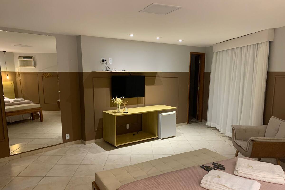 A hotel room with a bed, a desk with a TV, a small fridge, a chair, and a bathroom visible in the mirror.
