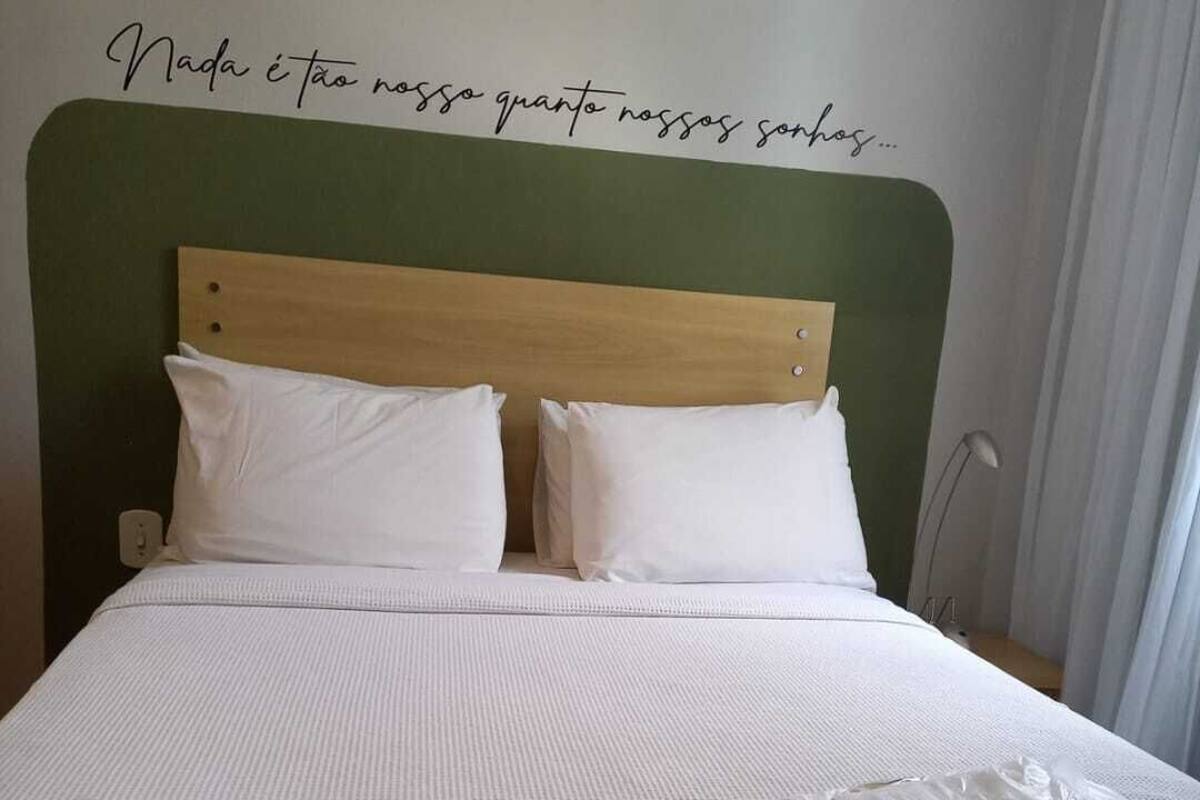 A neatly made bed with white bedding and pillows, a wooden headboard, and a wall with a motivational quote. There are two plastic bags with items on the bed.