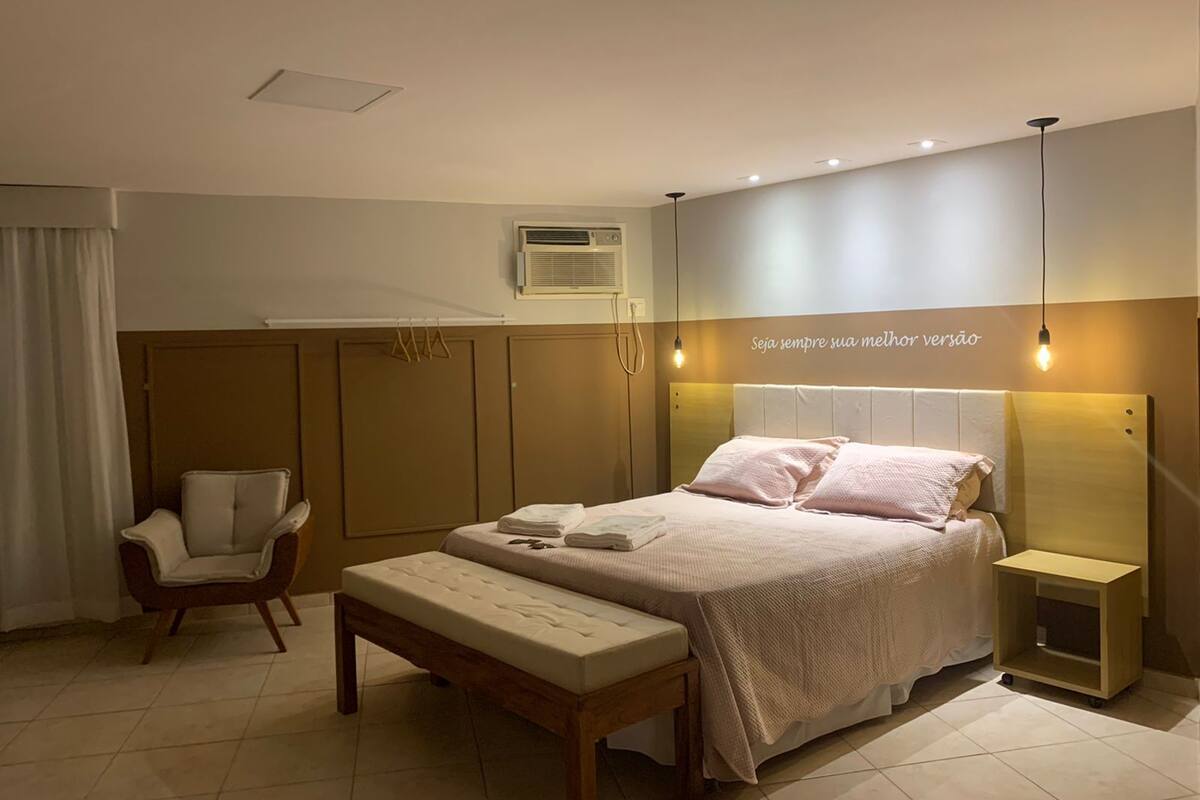 A bedroom with a bed, bedside tables, a chair, an air conditioning unit, and a wall-mounted light fixture.