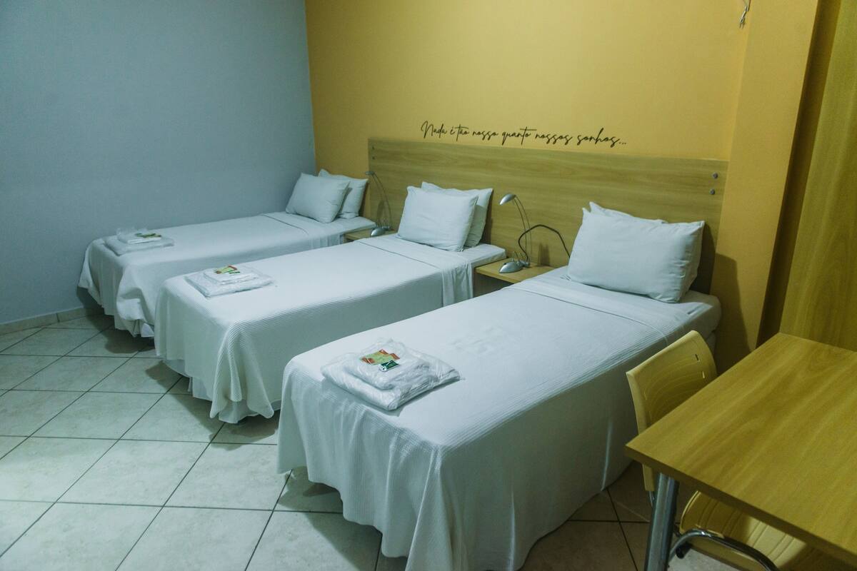 A hotel room with three single beds, each with a white bedspread and pillows, a wooden headboard with a motivational quote, a small wooden table, and a chair.
