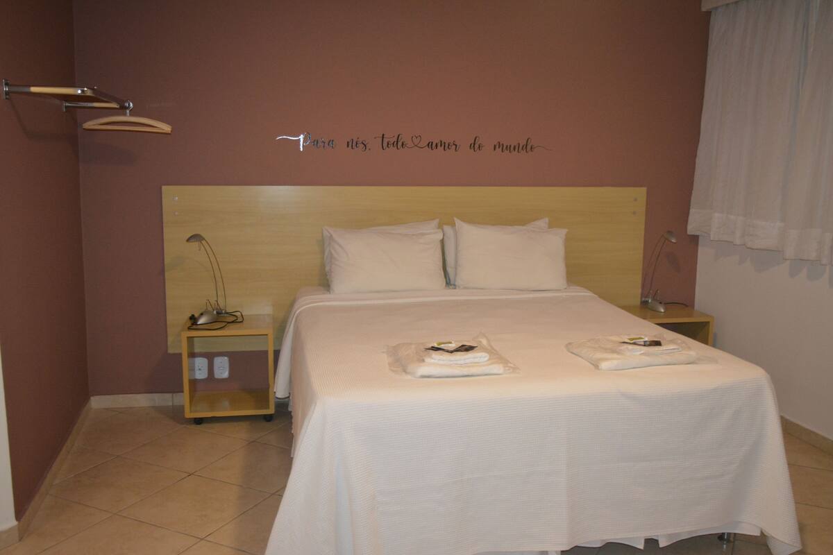 A hotel room with a bed, bedside tables, a wall-mounted lamp, and a sign with a message.