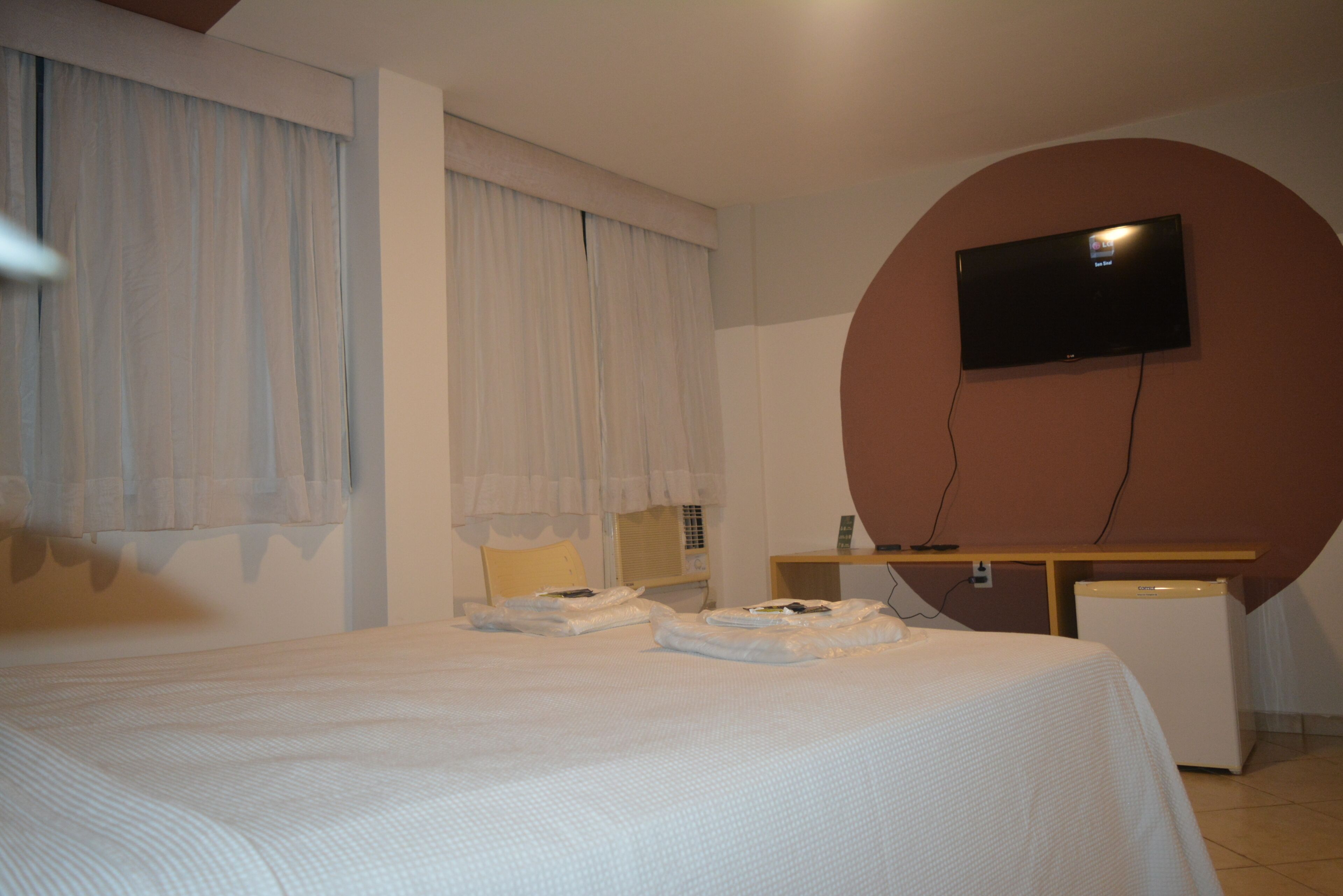 A hotel room with a bed, a television mounted on a wall, a desk, and a refrigerator.