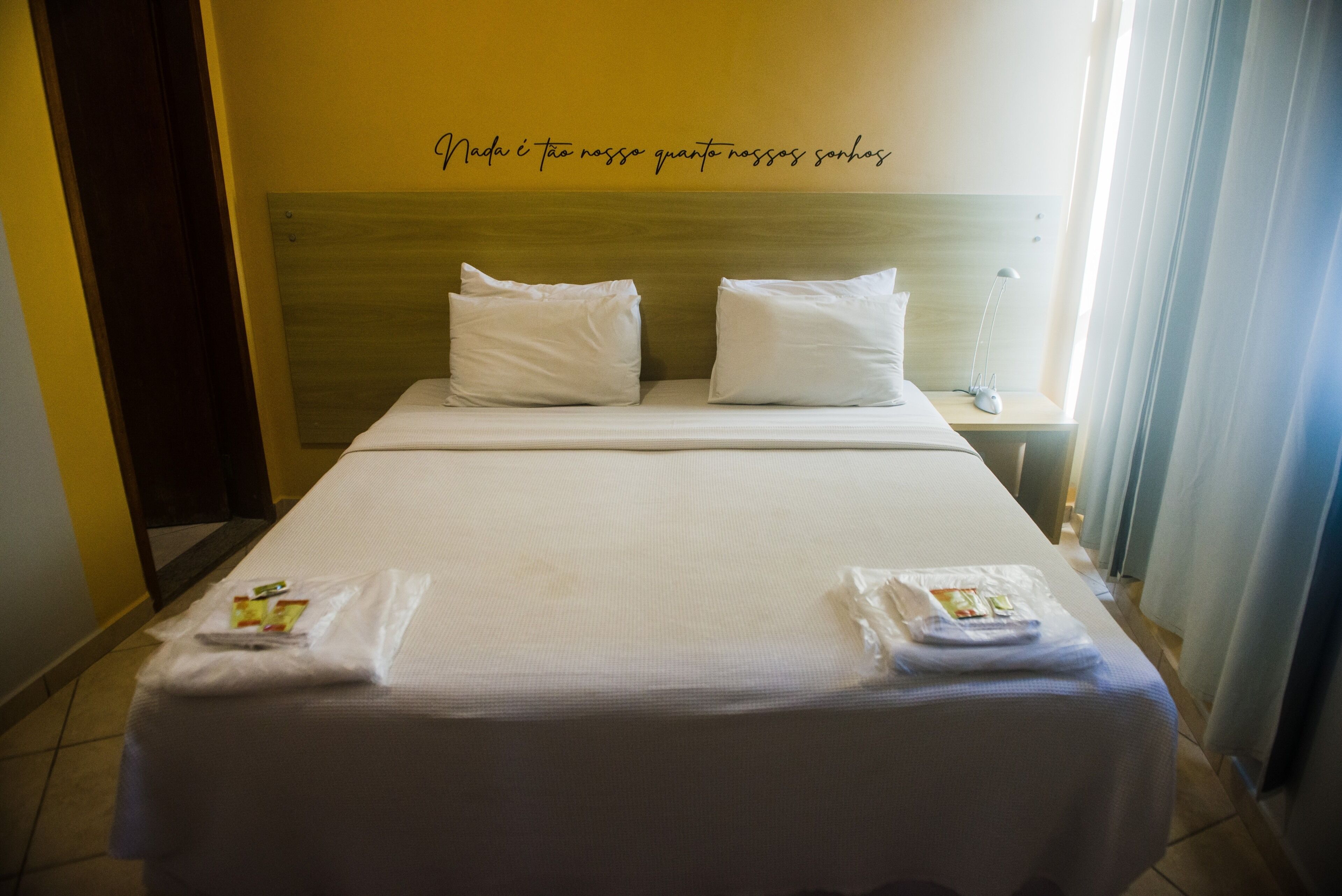 A hotel room with a neatly made bed, two pillows, a bedside table with a lamp, and a wall with a quote.