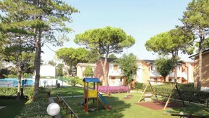 Ein Spielplatz mit Schaukeln, Rutschen und einer Kletteranlage, umgeben von Bäumen und einem Schwimmbad.