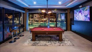 Una mesa de billar en una habitación con un ventanal grande, una zona de bar y una pantalla de televisión.