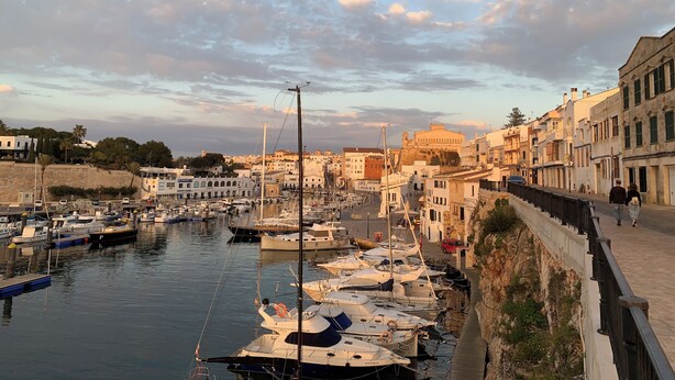 A marina with boats docked along a waterfront, buildings with balconies, and a stone wall.