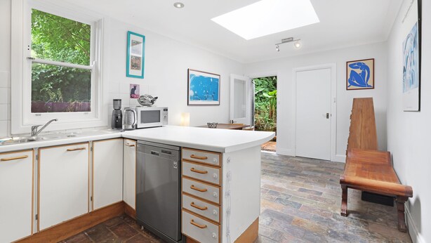 Una cocina moderna con armarios blancos, lavavajillas y una isla grande con cajones. Hay un banco de madera en la esquina y vistas a zonas verdes al exterior.
