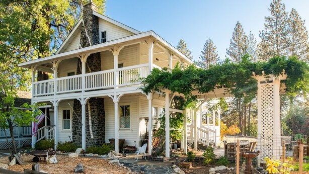 A two-story house with a porch, stone chimney, and a garden area with a wooden fence. - Inn at Sugar Pine Ranch (Groveland)