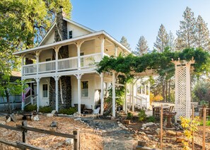 A two-story house with a porch, stone chimney, and a garden area with a wooden fence.
