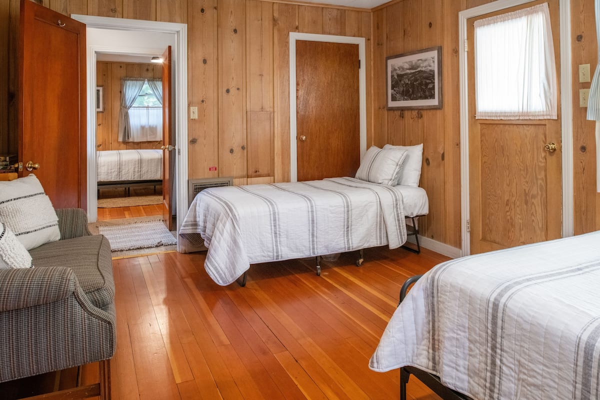 Una habitación con paredes y pisos de madera, dos camas con ropa blanca, una silla y una mesita.