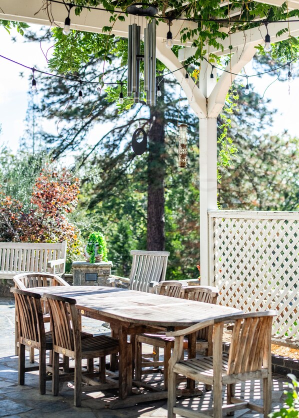 A wooden outdoor patio with a table and chairs, surrounded by a white lattice fence, under a pergola with hanging lights.