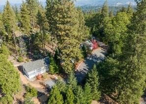 An aerial view of a residential area with a house surrounded by dense forest, a paved driveway, and a small parking area.