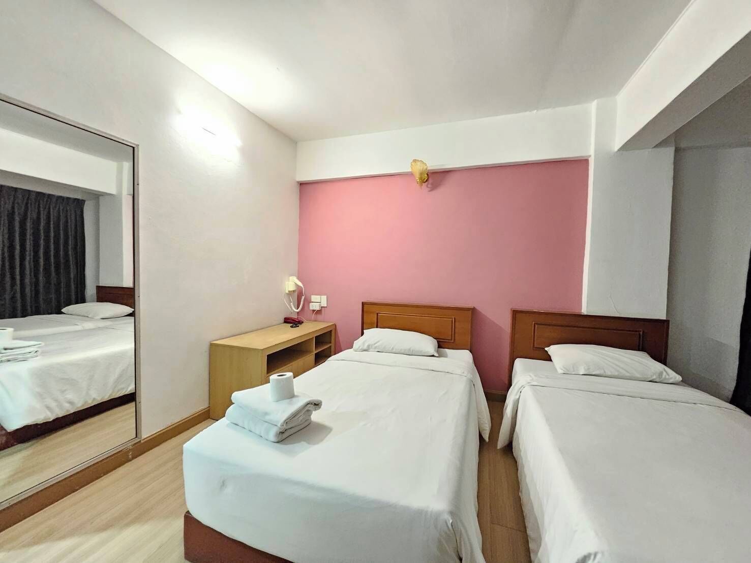 Une chambre d’hôtel avec deux lits simples, une table de chevet avec une lampe, un miroir et un mur d’accent de couleur rose.