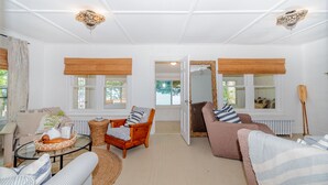 A spacious living room with a sofa, armchair, and coffee table. There are two windows with wooden blinds, a door leading outside, and a ceiling light fixture. - The Congamond Cottages: Private Lakefront Cottage (Suffield)