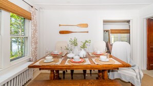 A dining room with a wooden table set for four, white walls, and a view of trees through a window with a leaf-patterned curtain. - The Congamond Cottages: Private Lakefront Cottage (Suffield)