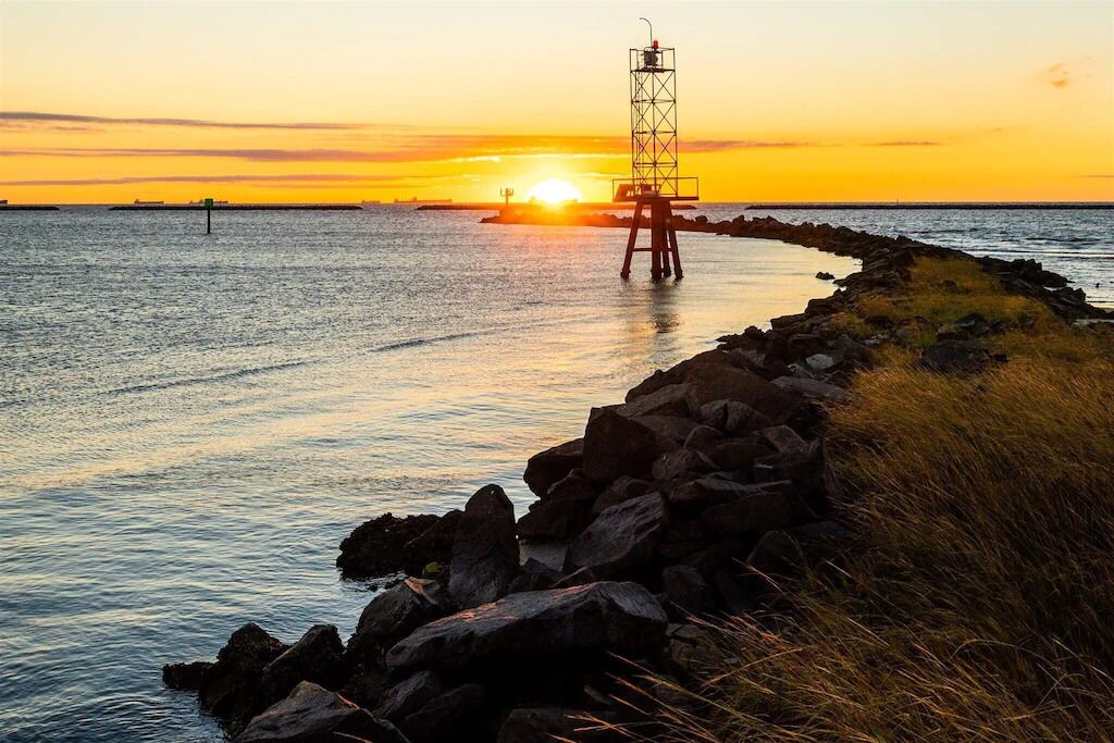 Eine Küstenlandschaft mit einem Leuchtturm bei Sonnenuntergang, felsigem Ufer und ruhiger See.