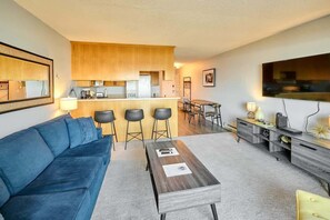 A living room with a sofa, a coffee table, a kitchen area with a bar, and a TV mounted on the wall. - Updated Oceanfront Condo -Ground Floor- Fireplace! (Lincoln City)