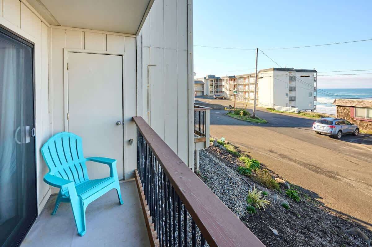 Een balkon met een blauwe stoel, uitzicht op een straat met gebouwen en een auto, en een heldere lucht.