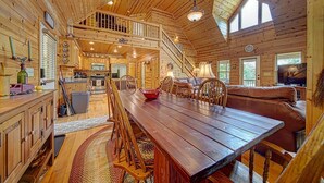 Un interior espacioso de cabaña de troncos con una gran mesa de comedor, muebles de madera, una escalera que lleva a un altillo y una sala de estar acogedora con un sofá y televisor.