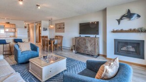 A living room with a sofa, coffee table, and a fireplace. There is a television mounted on the wall and a dining area with chairs. - Ocean View Condo - Second Floor - Pet Friendly! (Lincoln City)