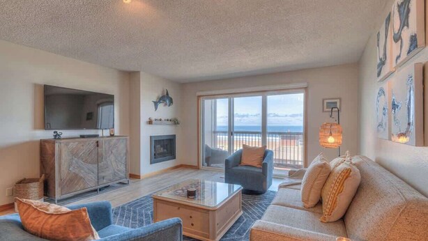 A living room with a sofa, armchair, coffee table, TV, and a view through large windows. - Ocean View Condo - Second Floor - Pet Friendly! (Lincoln City)