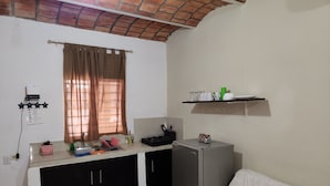 A compact kitchen with a refrigerator, a sink, and a countertop with various items. There is a window with curtains, a wall-mounted shelf with dishes, and a white sofa with a patterned pillow. - Coral House II (Sayulita)