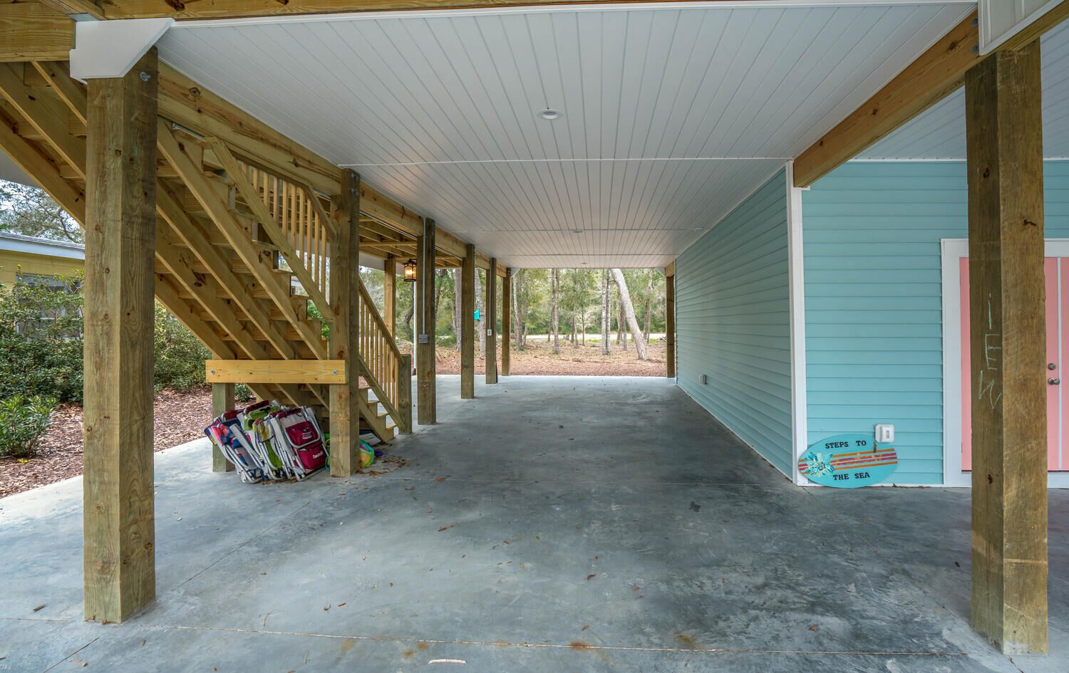 Een overdekt terras met houten balken en een betonvloer, dat leidt naar een huis met een blauwgroene gevel en een roze deur.
