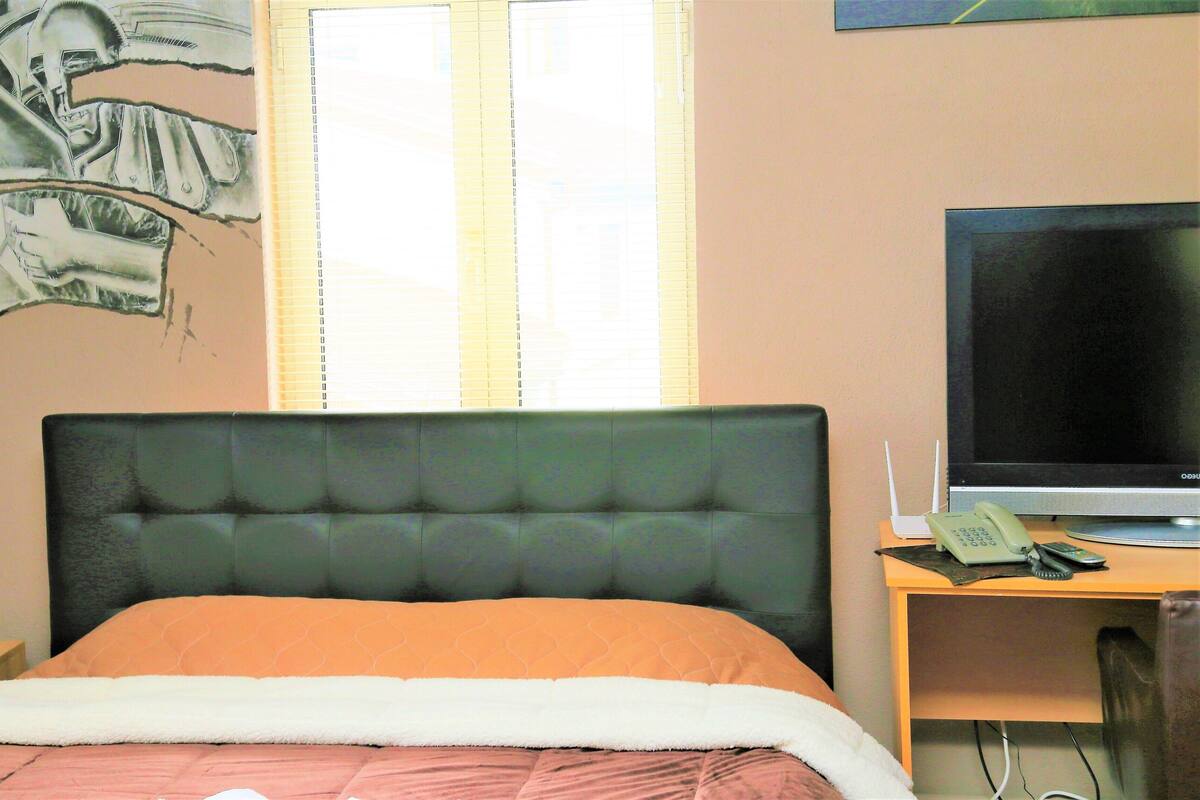 A hotel room with a bed, a desk with a computer and phone, and a window with blinds.
