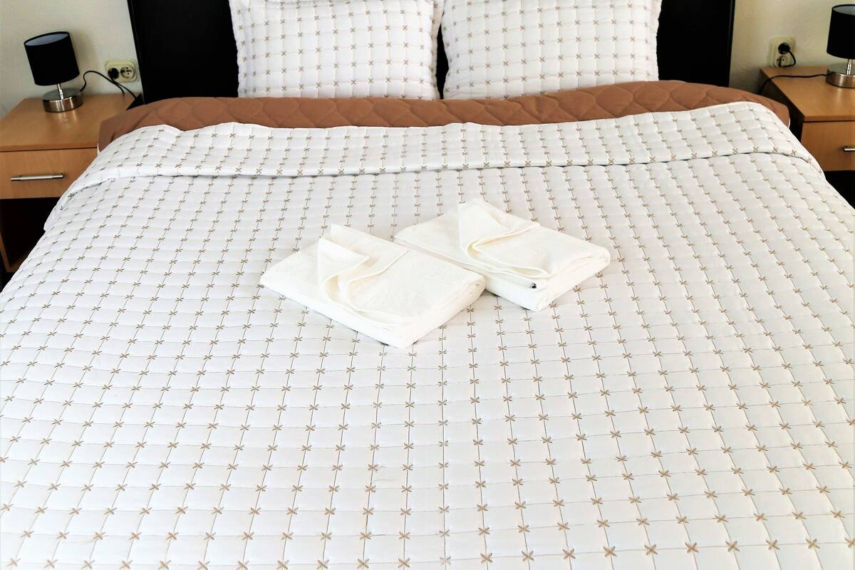 A neatly made bed with a white duvet and two pillows, a wooden nightstand with a lamp, and a wall socket.