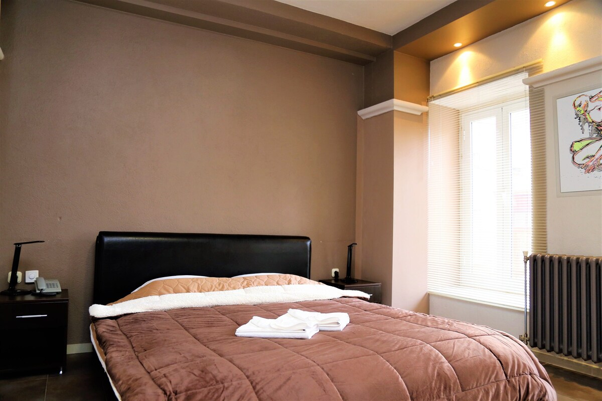 A neatly made bed with a brown quilt, a bedside table with a lamp, and a window with blinds.