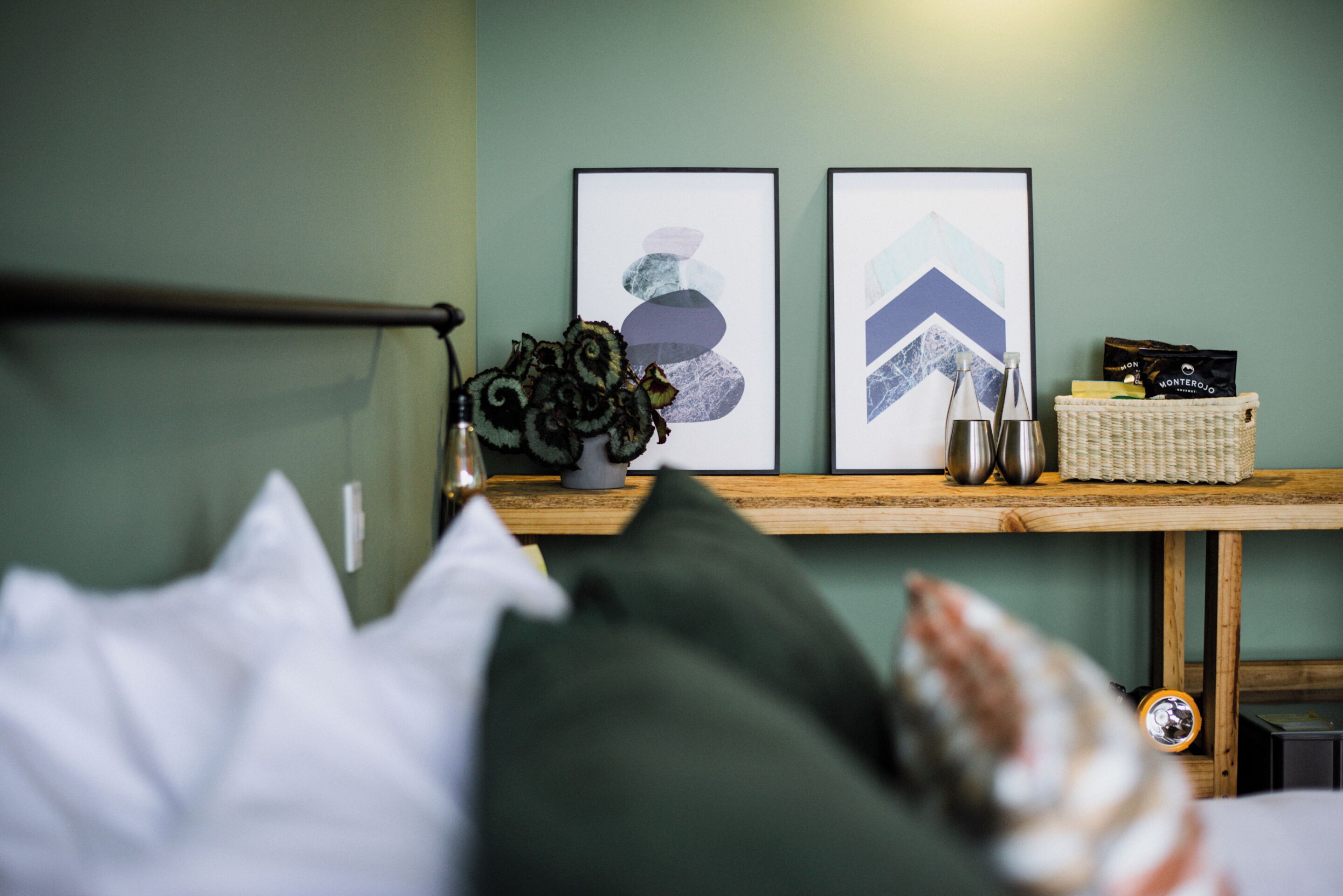 A bedroom with a bed, pillows, a bedside table with a vase of flowers, framed artwork on the wall, and a basket with items on the table.