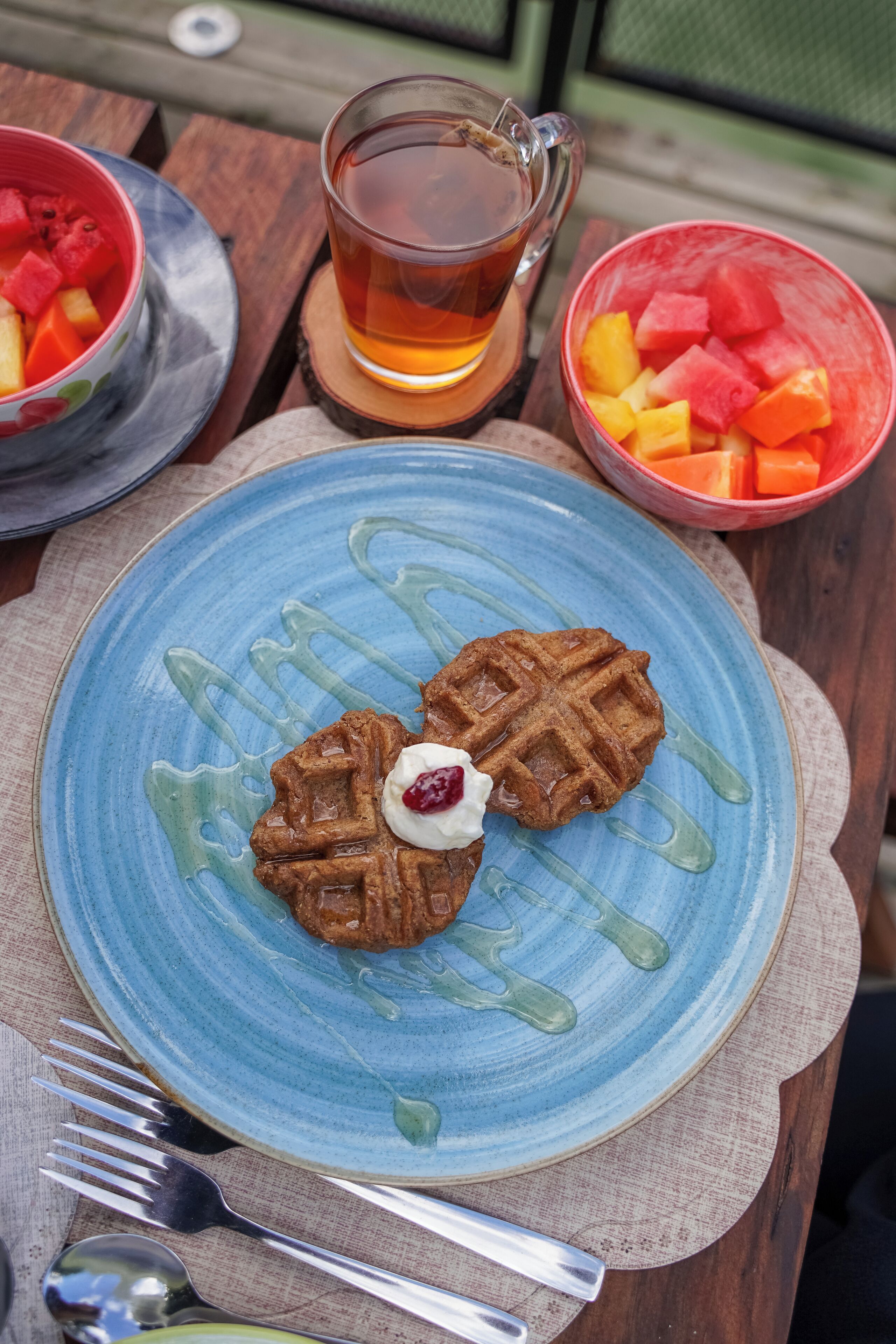 A table with a blue plate containing waffles and a glass of tea, accompanied by bowls of fruit.