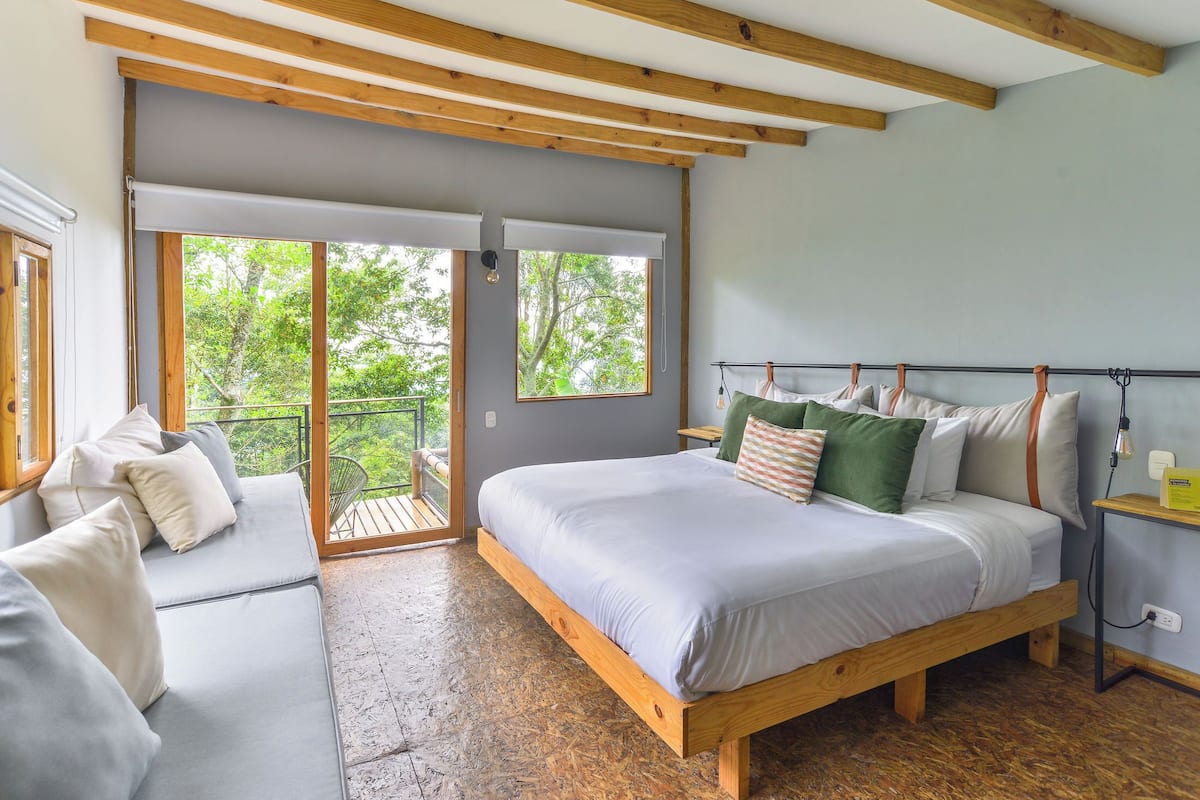 A bedroom with a large bed, a sofa, and a balcony with a view of trees.