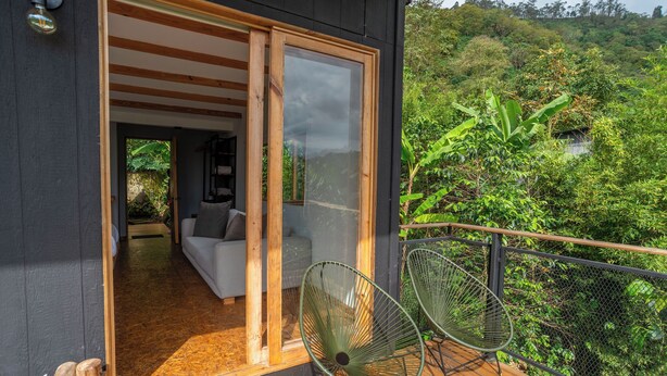 A modern house with a wooden deck, a glass door leading to a living area with a sofa and chairs, and a lush green forest view. - Hotel La Palma y El Tucan - Coffee & Nature (Zipacón)